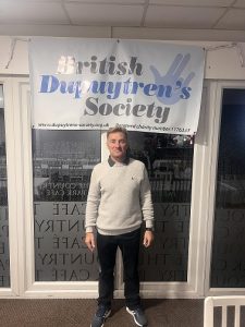 Man stands in front of a large window, with a banner behind him that reads 'British Dupuytren's Society'